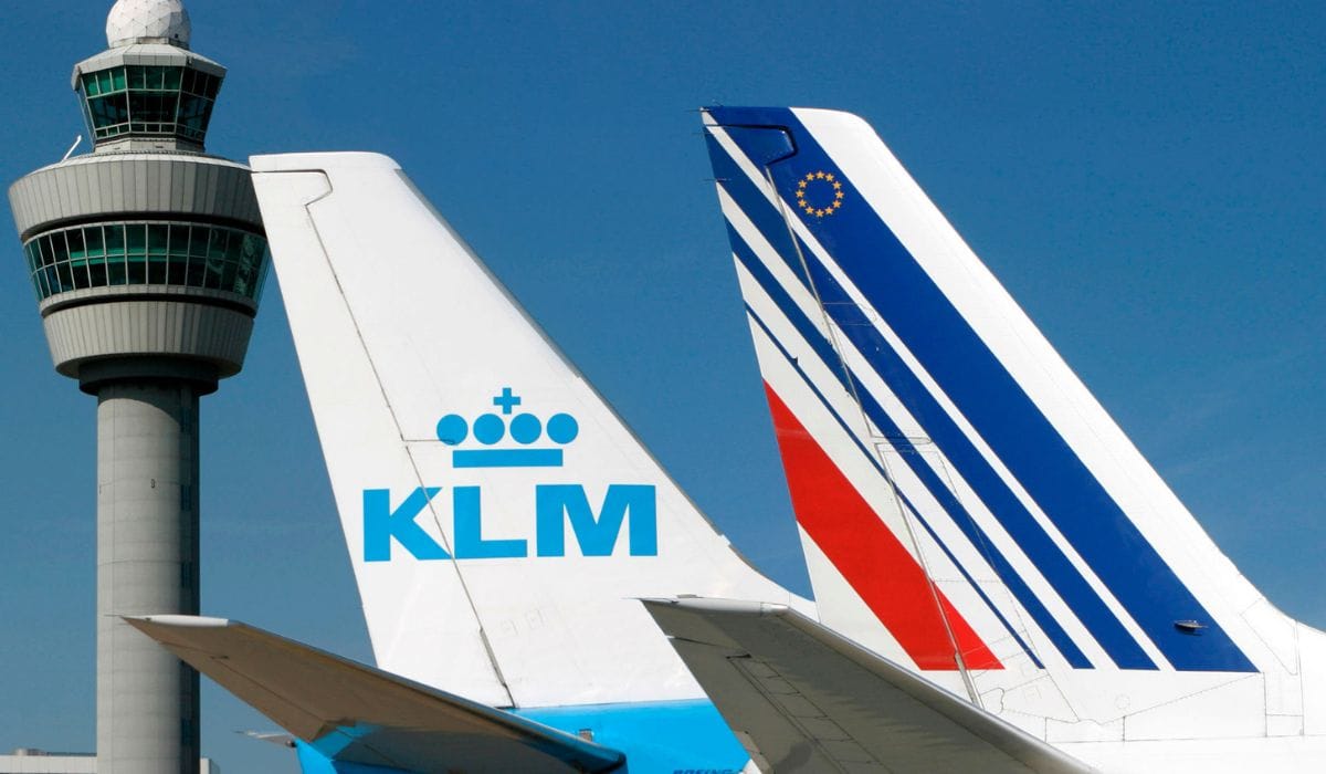 The tail fins of a KLM Royal Dutch Airlines plane and an Air France aircraft parked side-by-side on an airport tarmac. In the background, the iconic air traffic control tower of Amsterdam Airport Schiphol stands against a clear blue sky.